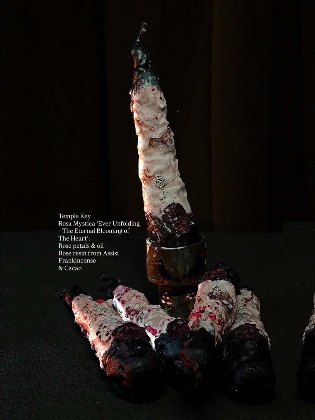 Art installation titled 'Ever Unfolding - The Eternal Blooming of The Heart' featuring a tall, textured sculpture resembling a burning candle, with a smoky black tip, surrounded by smaller similar sculptures, all on a black background. The sculpture 