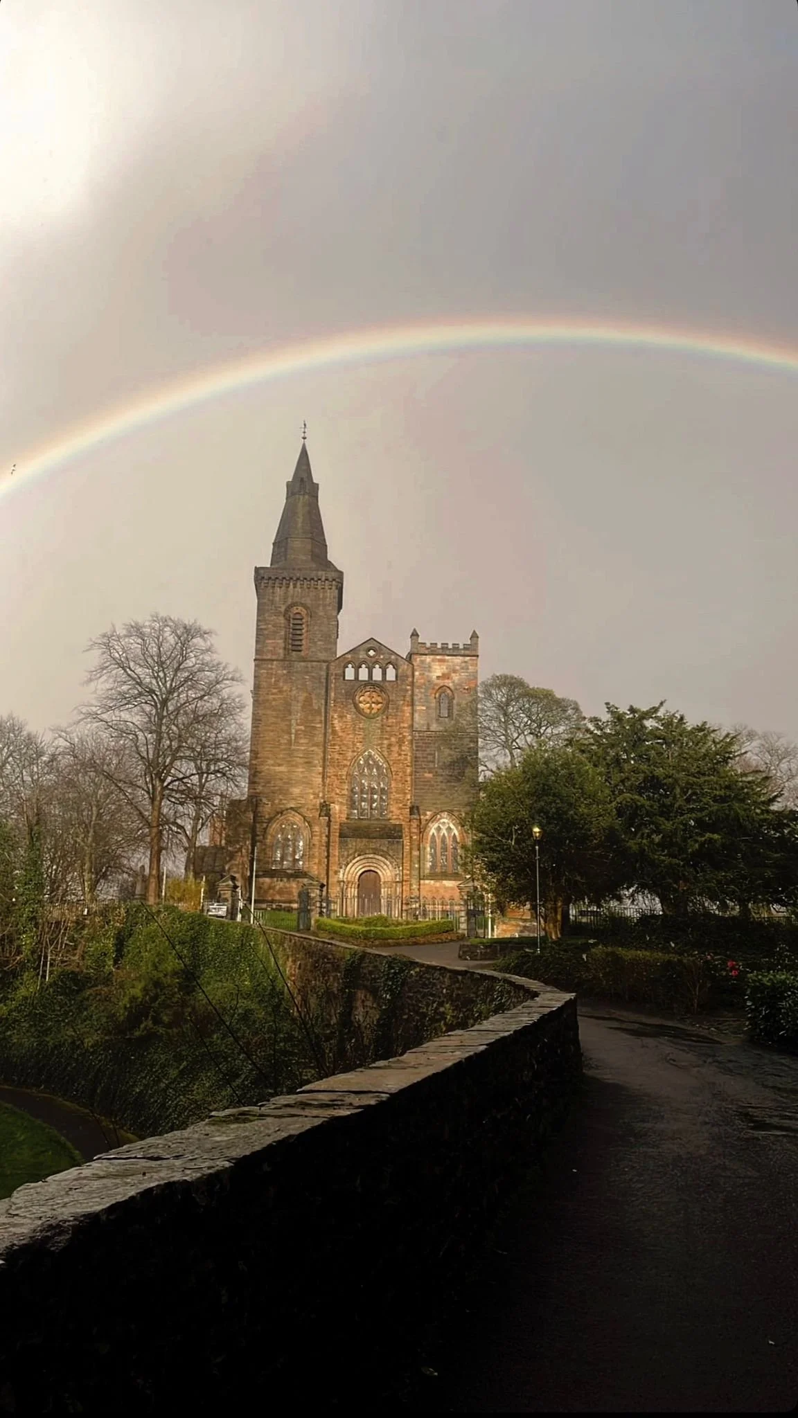 A church with a tall steeple underneath a faint rainbow on a cloudy day.