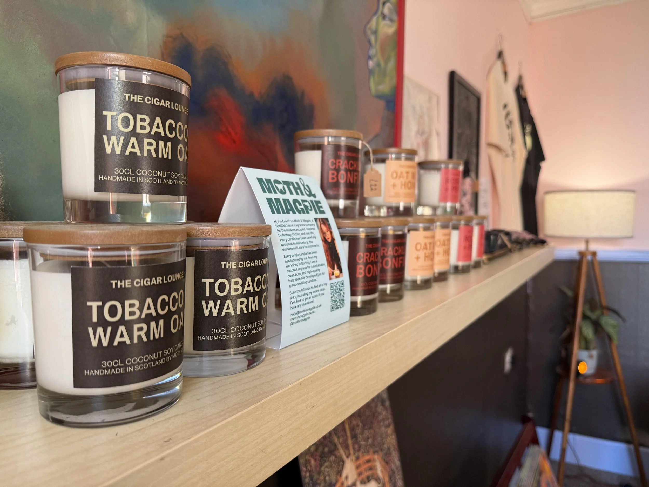 Display of candles with labels such as 'Tobacco Warm Oud' and 'Crackle Bonfire' on a wooden shelf in a cozy room with colorful artwork and a standing lamp.