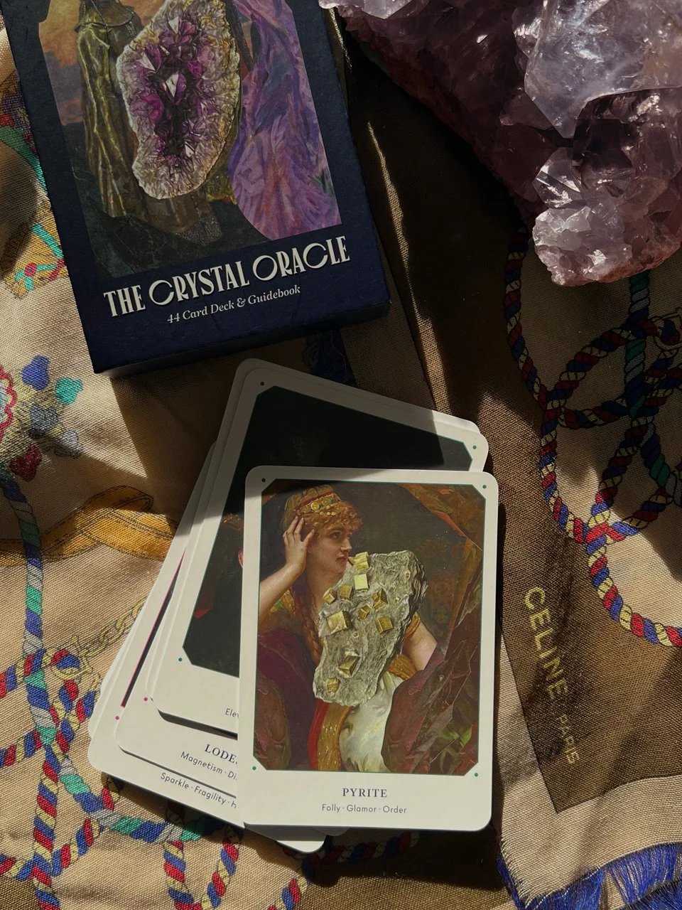 A set of tarot or oracle cards spread out on a cloth, with a box labeled 'The Crystal Oracle' 44 card deck and guidebook at the top, and a large amethyst crystal to the right.