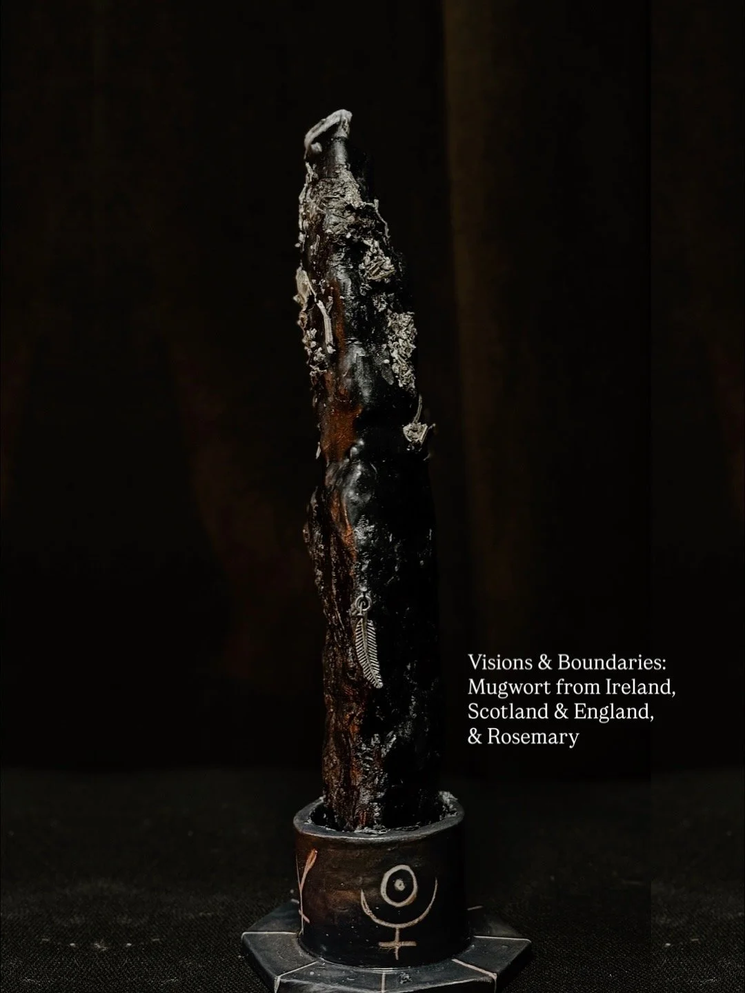 A charred mugwort stick with a necklace lying beside it, placed in a small cup with a female symbol and a spiral design. The background is black, and a caption mentions "Visions & Boundaries: Mugwort from Ireland, Scotland & England, & Rosemary."