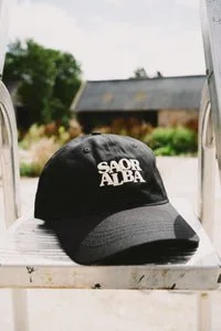 Black baseball cap with white text reading 'Saor Alba' resting on a white wooden surface outdoors.