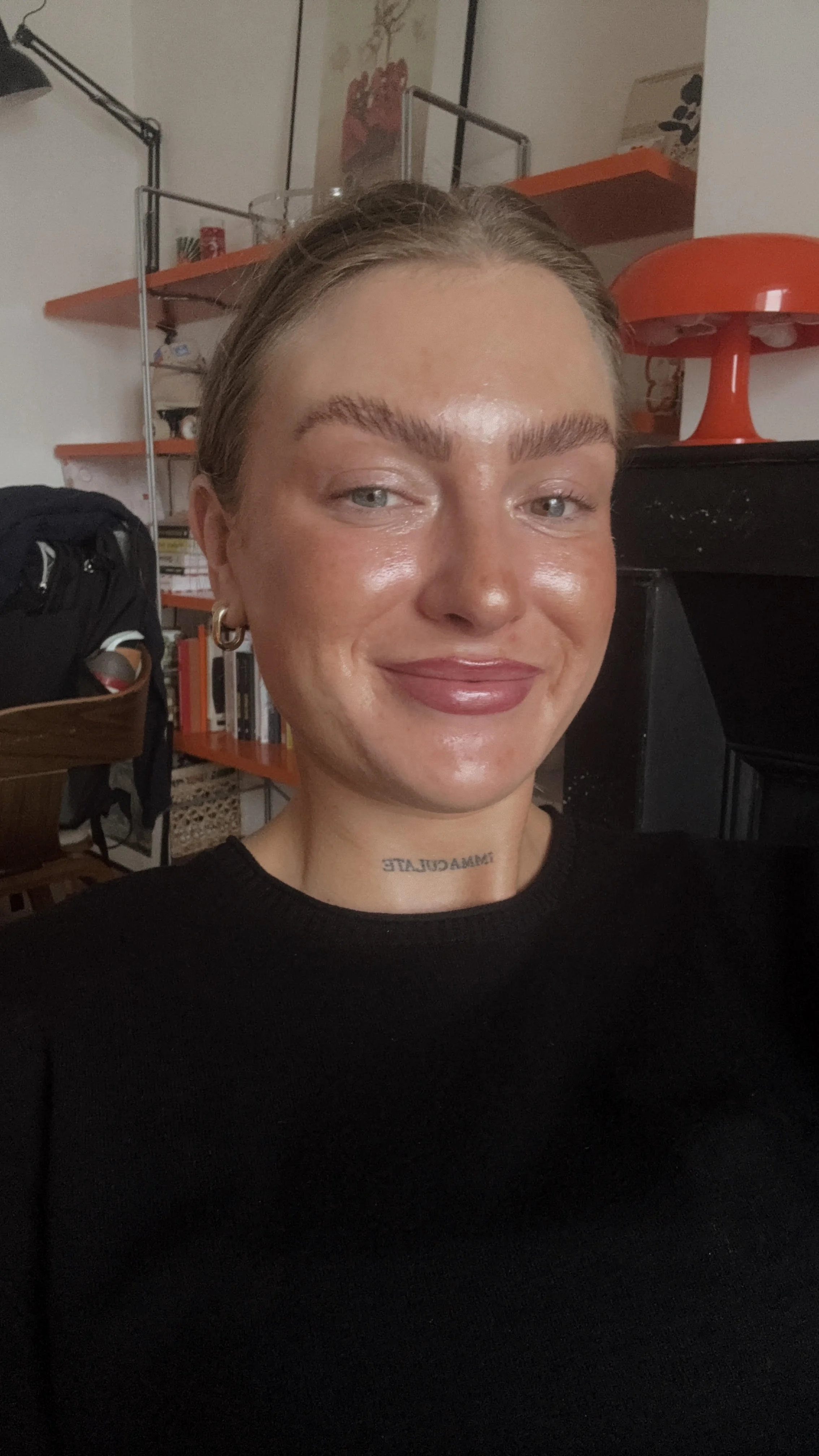 Close-up of a woman with short blonde hair, wearing a black top and gold hoop earrings, smiling indoors with a bookshelf, a red lamp, and artwork in the background.