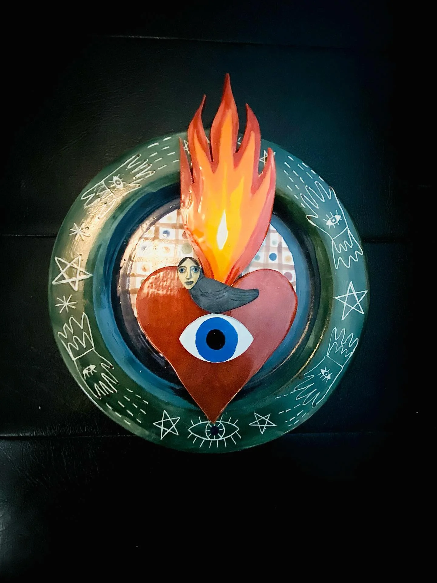 Decorative circular glass piece featuring a red heart with an eye, a face, a bird, and a flame above, surrounded by mystical symbols and drawings.