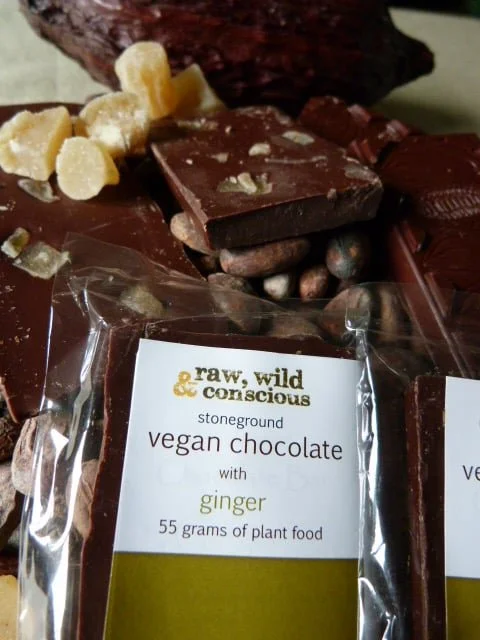 A package of raw, wild, conscious vegan chocolate with ginger, labeled as 55 grams of plant food, placed on a surface with broken pieces of chocolate and vanilla beans in the background.