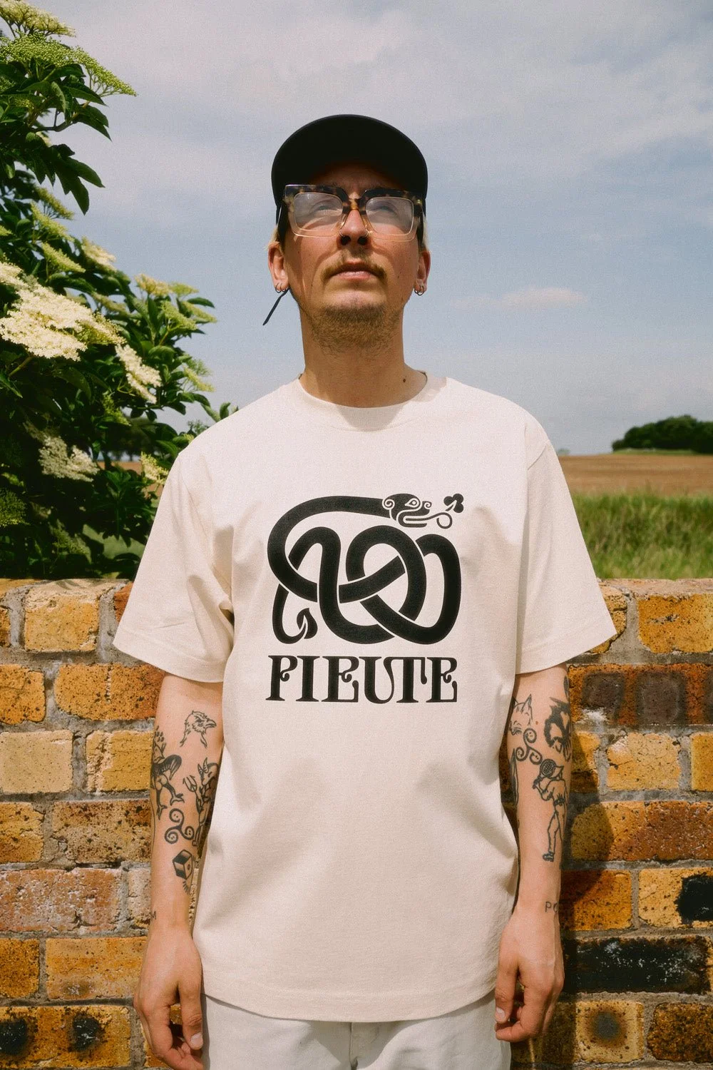 A man wearing a white T-shirt with a black knot design and the word "PIETTE" on it, standing outdoors in front of a brick wall with greenery and a field in the background, under a partly cloudy sky.