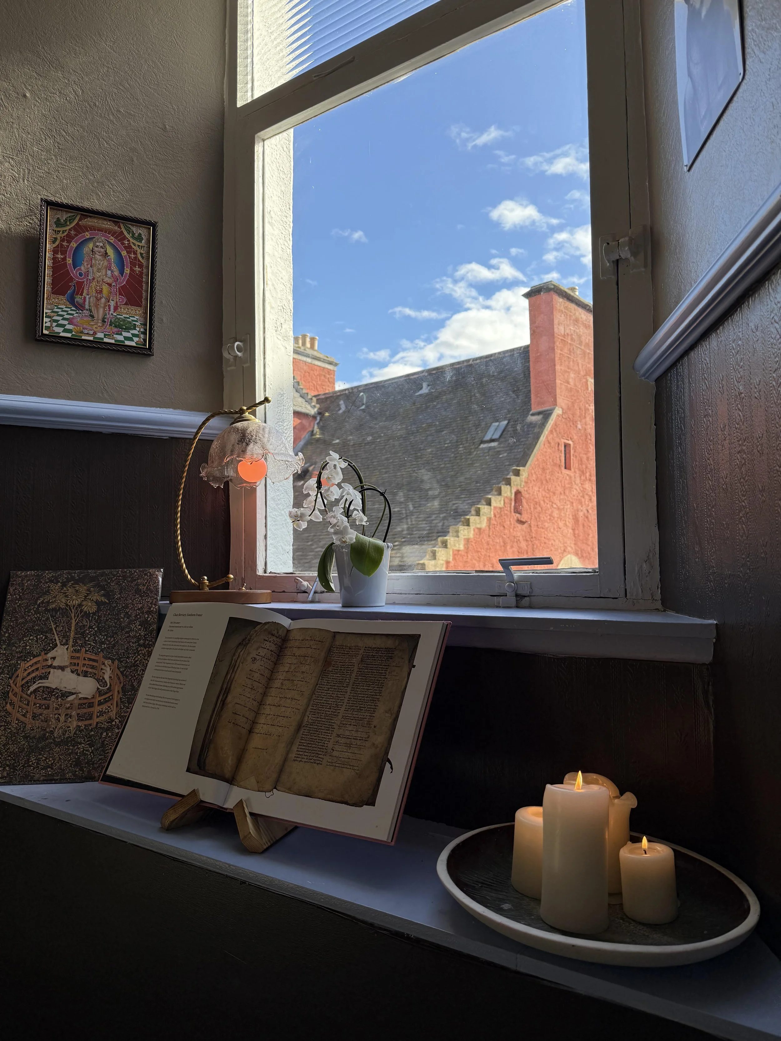 A windowsill with a white orchid in a pot, an open book on a stand showing an ancient manuscript, a candle holder with three lit candles, and a photo of a deity on the wall. Outside the window, there is a view of a red brick building roof and a blue sky with clouds.