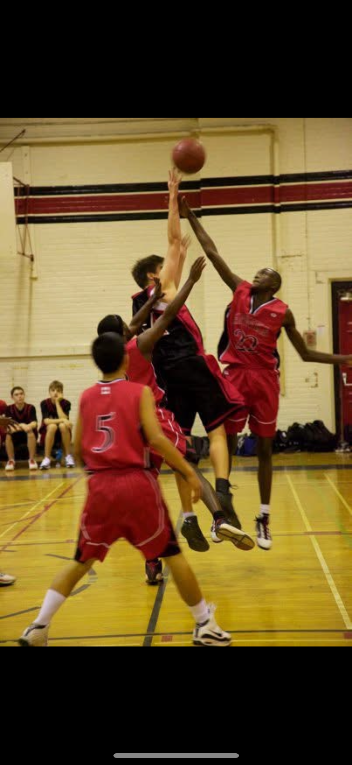 A basketball game with players in red jerseys jumping to block a player in a black and red jersey attempting a shot, with seated players watching in the background.