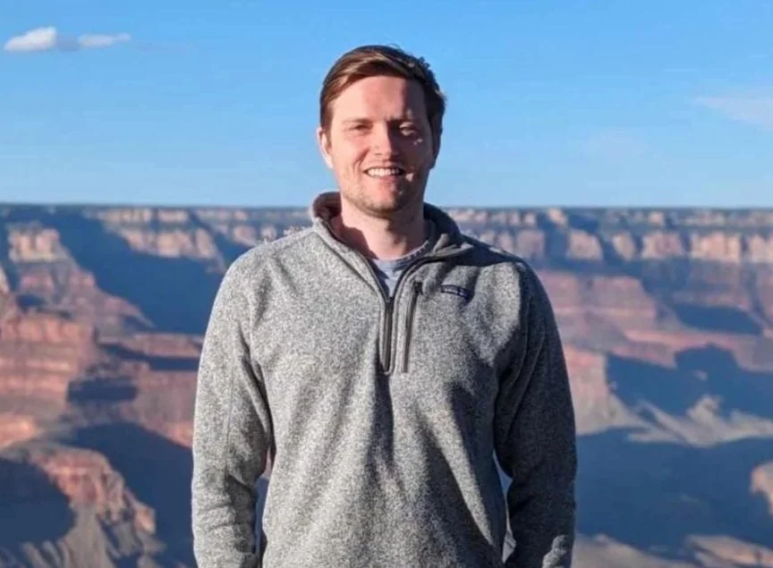 A man standing smiling in front of the Grand Canyon during daytime, wearing a gray zip-up jacket and a blue sky background.