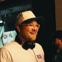 A man with glasses, a cap, and headphones around his neck smiling in a dimly lit environment