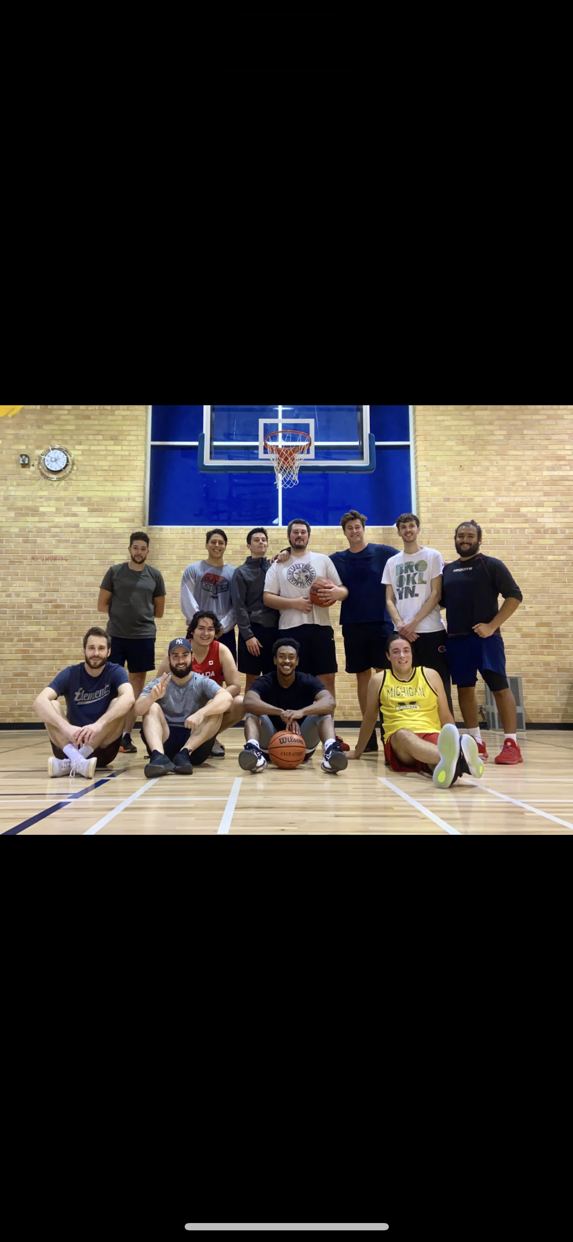 Group of basketball players in a gymnasium, posing for a photo in front of a basketball hoop and a brick wall, with some sitting and others standing, holding basketballs.