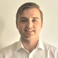 Portrait of a young man with light brown hair, wearing a white collared shirt, smiling against a neutral background.