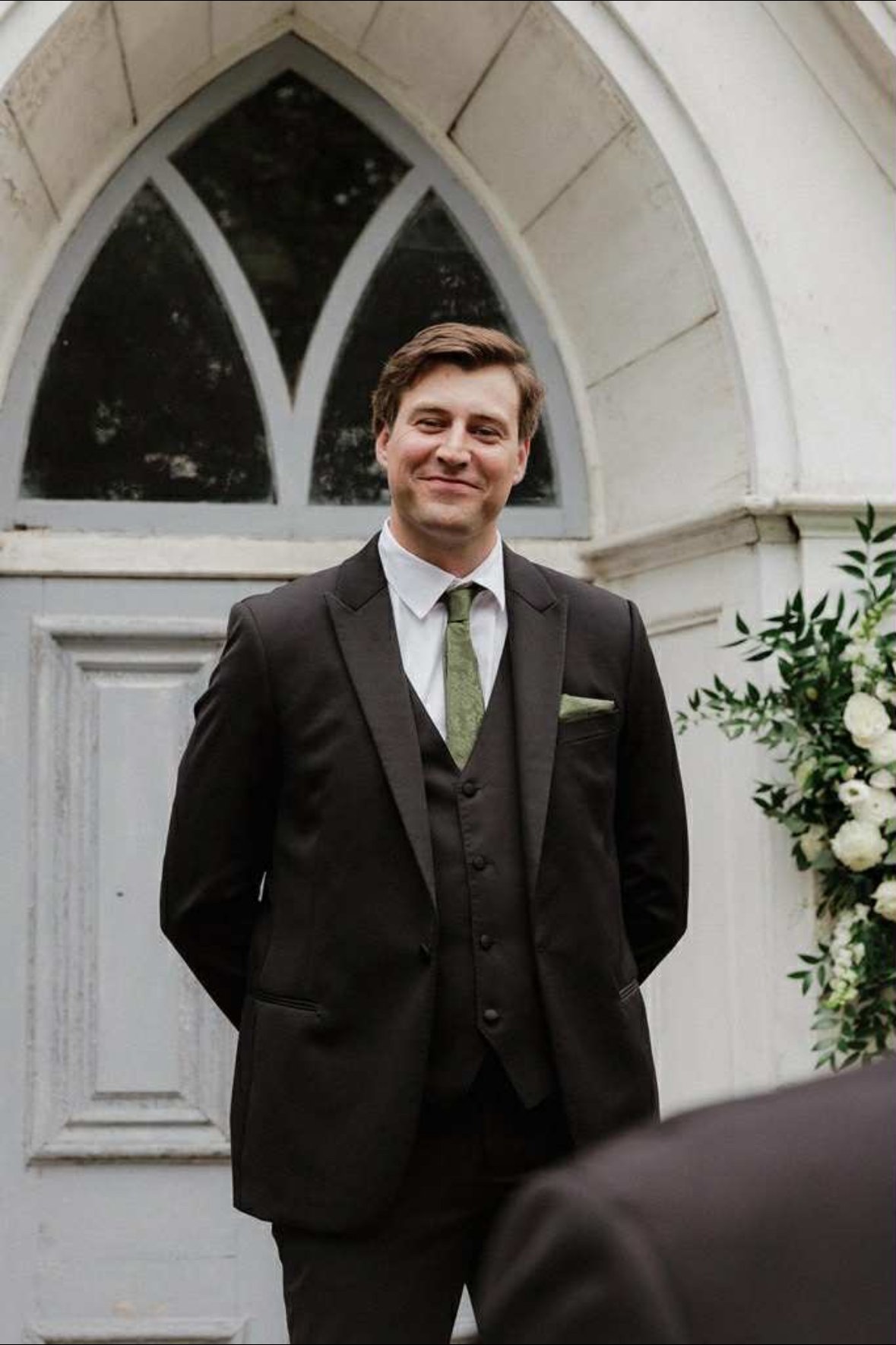 A man in a black suit smiling while standing outside near a Gothic-style arched window and white wall, with a floral arrangement nearby.
