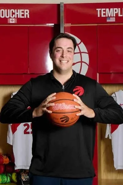 A man in a black Toronto Raptors hoodie holding a basketball, standing in front of red lockers with the words 'Boucher' and 'Temple' and a Toronto Raptors jersey with number 17 hanging.
