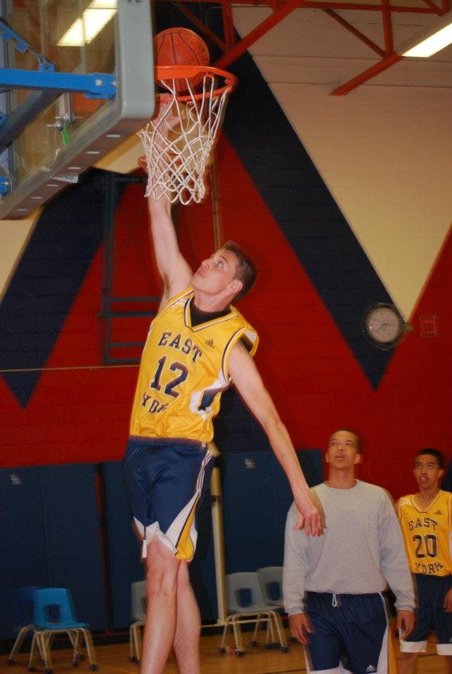 A basketball player in yellow and blue jersey number 12 dunking the ball into the hoop during practice or game, with two other players watching in the background.
