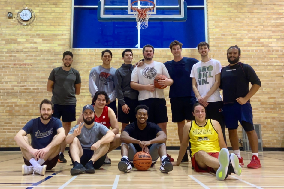Group of young adults in a gym posing with basketballs in front of a basketball hoop and brick wall.
