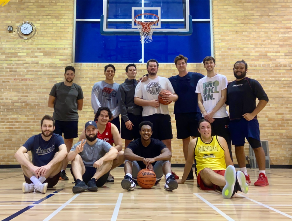 A group of young adults on an indoor basketball court, some sitting and others standing, with a basketball hoop behind them