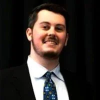 A young man with dark hair and a beard smiling in front of a dark curtain, wearing a suit and tie.