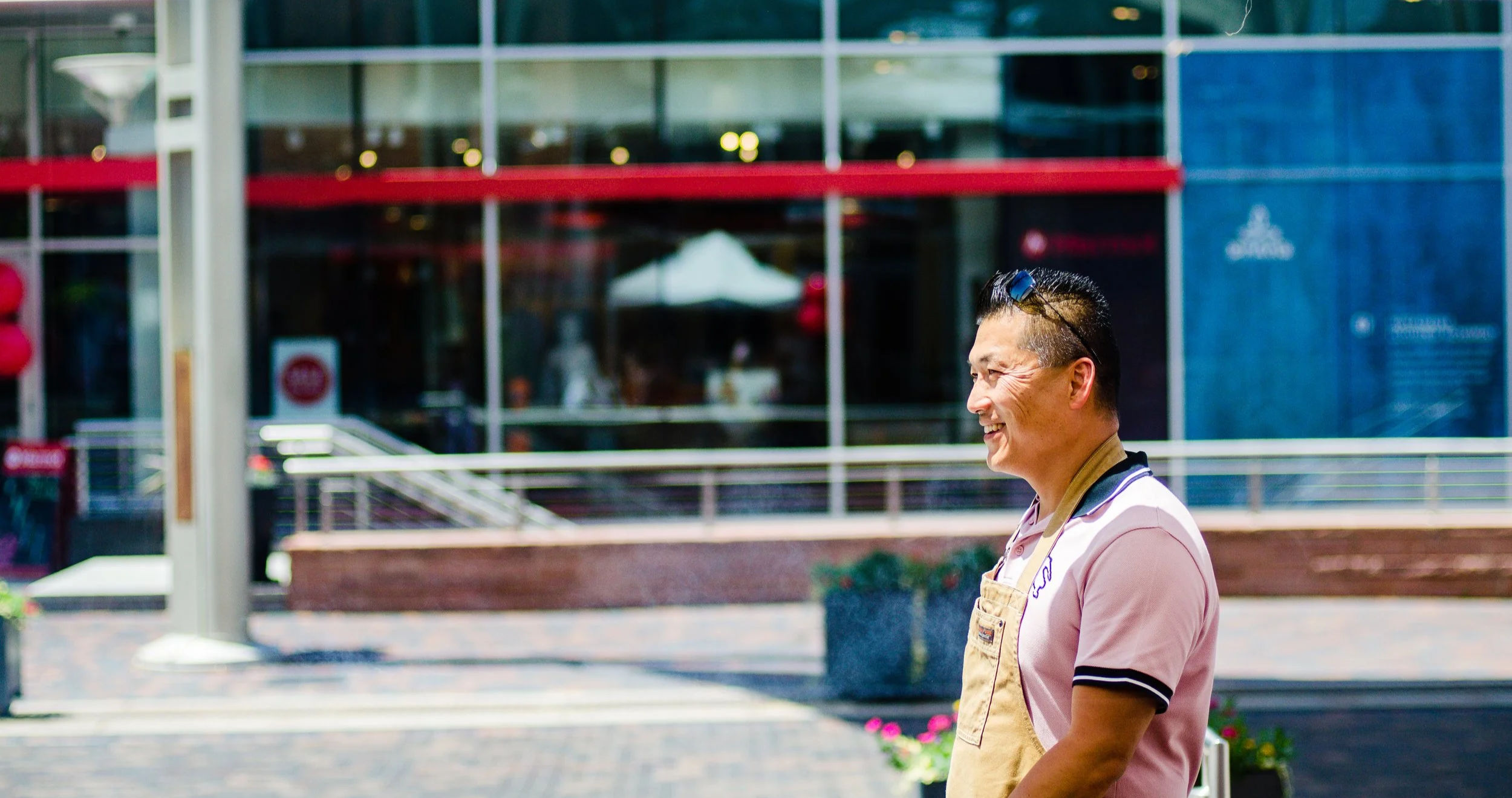 A smiling man wearing a pink polo shirt and an apron standing outdoors in an urban area, with a modern glass building and some red decorations in the background.