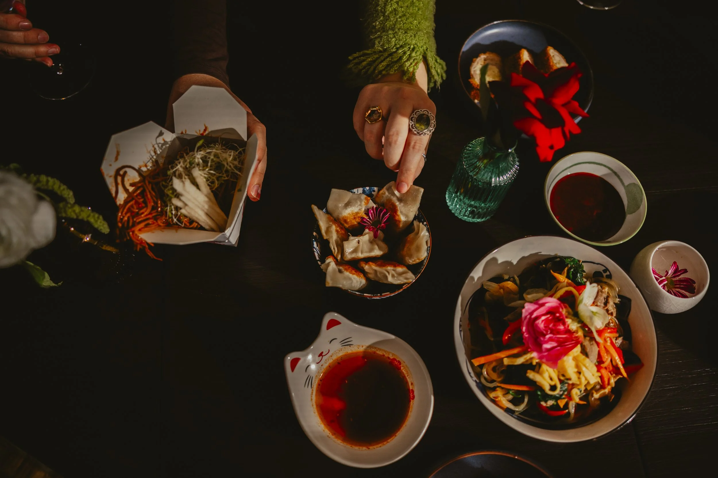 Hand reaching for dumplings on a black plate surrounded by various Asian dishes, including noodles, salad, and dipping sauces, on a dark table with a red flower in a glass vase.
