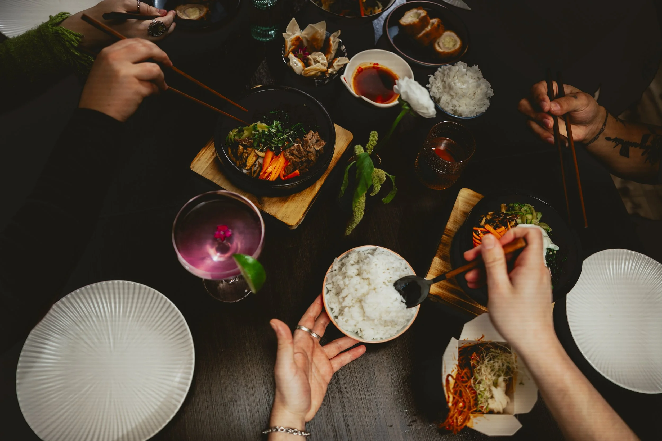 People at a dinner table with various Asian dishes including rice, sushi, and vegetables, along with drinks and a flower centerpiece.