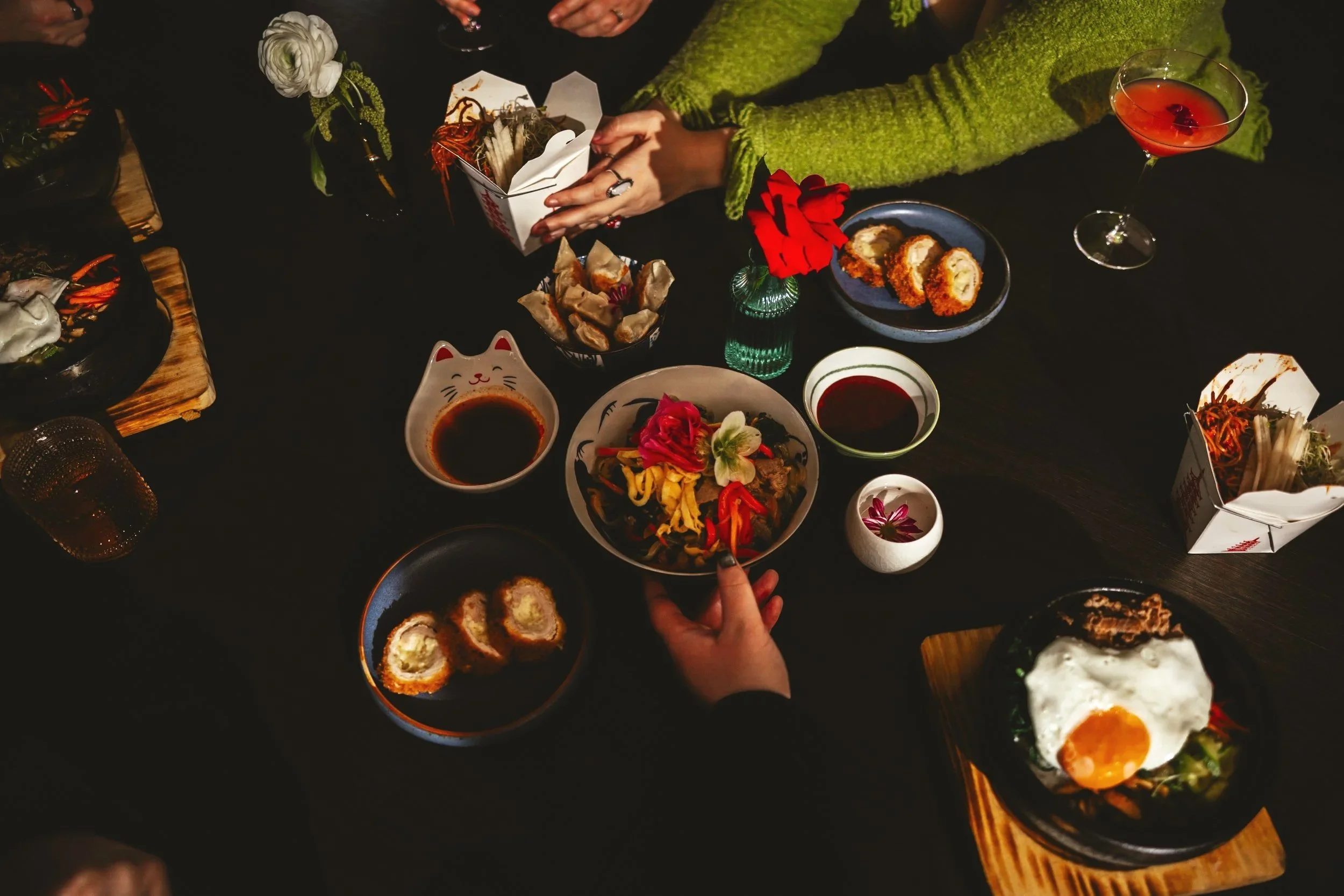 A table with various dishes including sushi, spring rolls, salad, fried rolls, and drinks, with people reaching for food. The table is decorated with flowers and a black tablecloth.