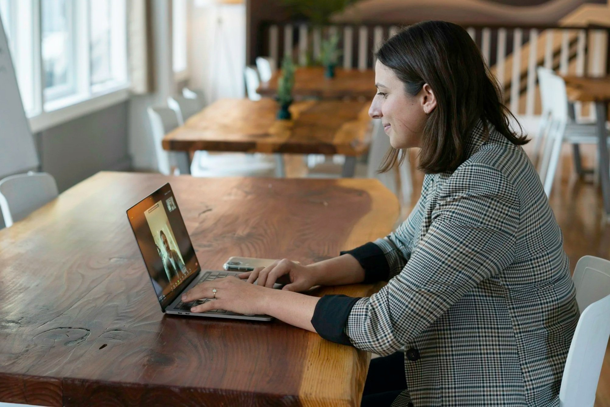Woman in checkered blazer speaking via video call on laptop at wooden table in a bright room.