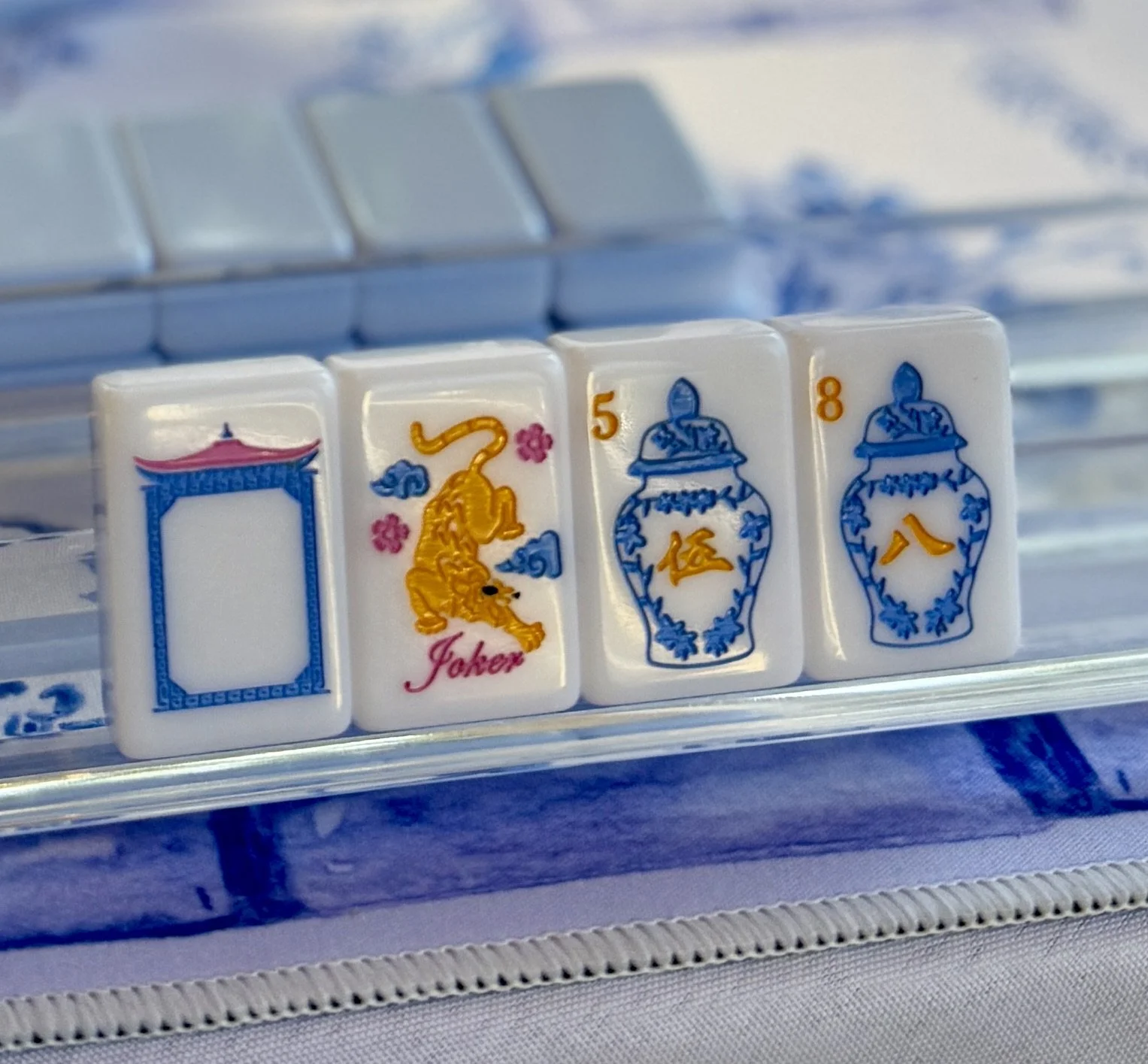 Close-up of Mahjong tiles, including a blank tile, a joker tile with a tiger illustration, and numbered blue and white vases, on a clear plastic tray.