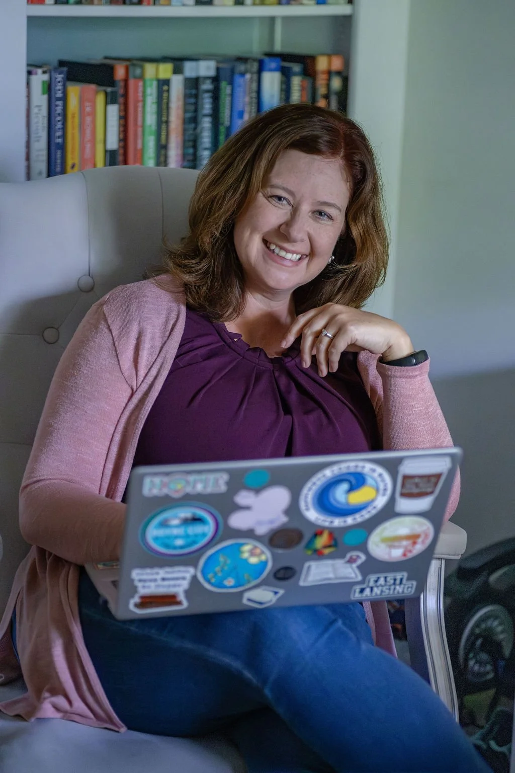 Fiction book coach, Miranda Keskes, is smiling, sitting in a chair, wearing a purple top and pink cardigan, with a laptop in front of her decorated with various stickers and a bookshelf filled with books in the background.