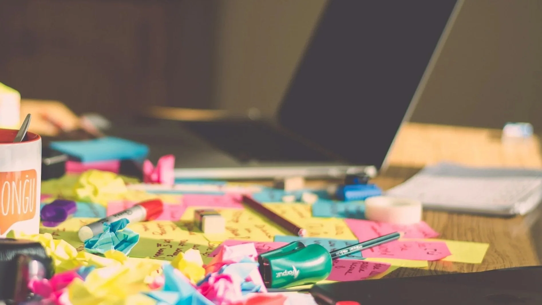 A teacher-mom’s cluttered desk with a closed laptop, open notebook, kid items, graded papers, and a half-finished coffee, showing interrupted writing time.