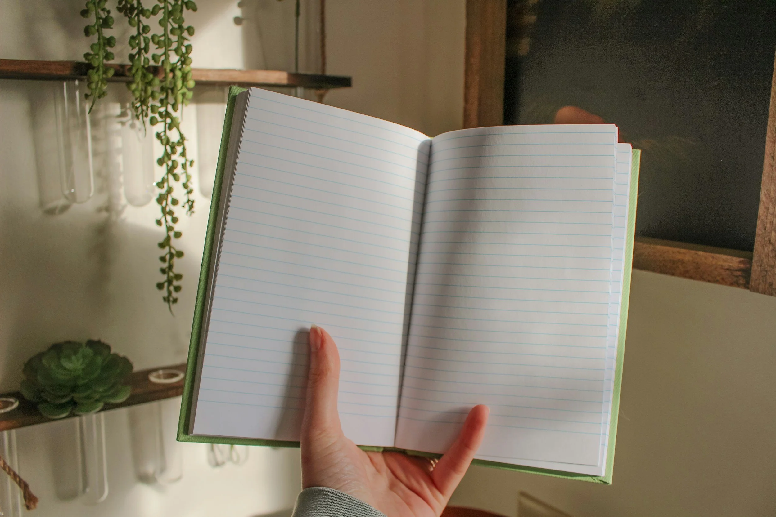 Open notebook with lined pages being held in a person's left hand. Decorative shelves with succulents and hanging plants in the background.