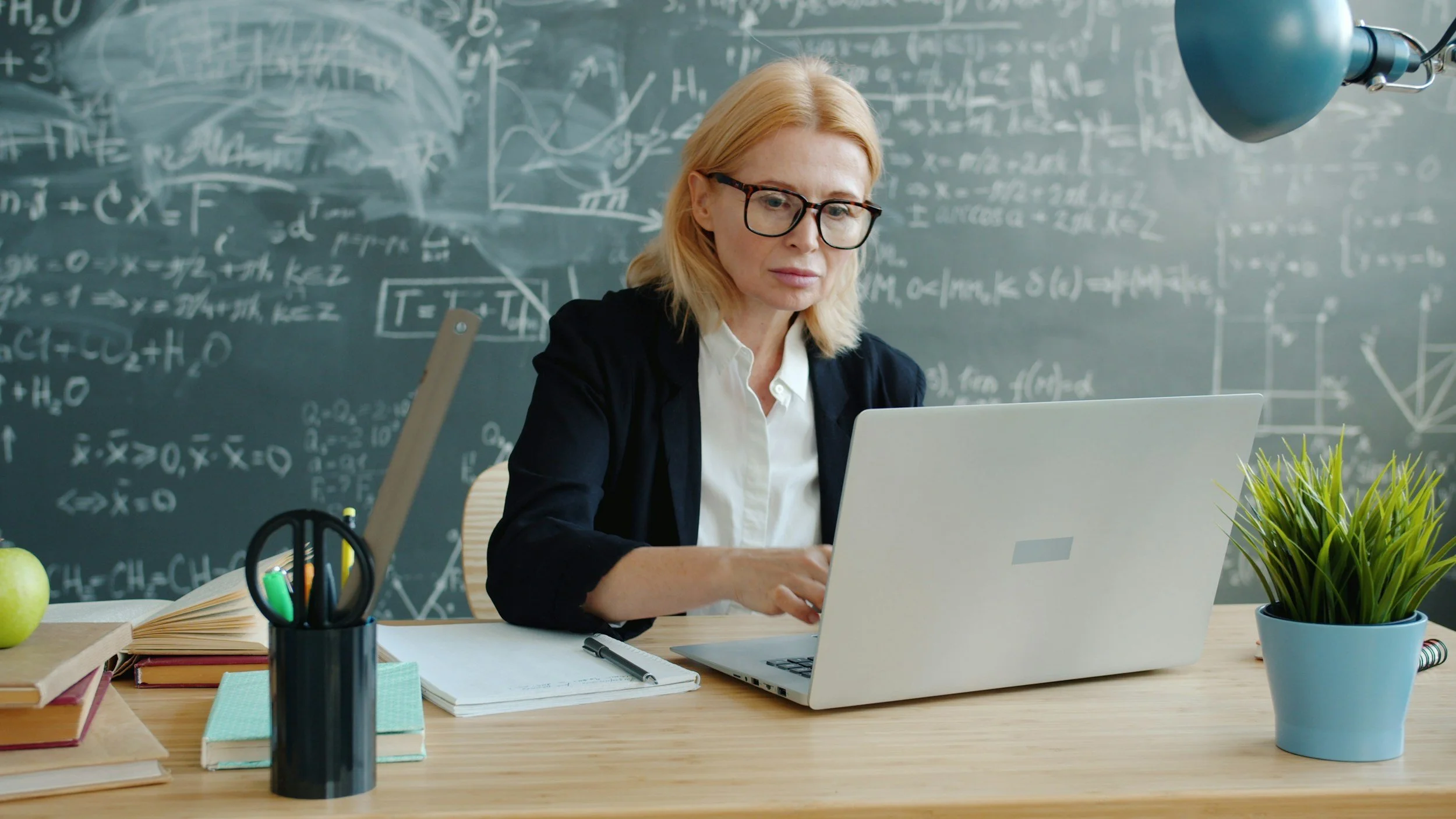 A blond woman with glasses typing on her laptop at her teacher's desk. A chalkboard is behind her. She looks calm and focused.
