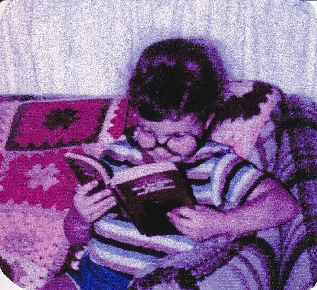 Picture of a little girl from the 1980s, wearing oversized glasses and reading an adult paperback.