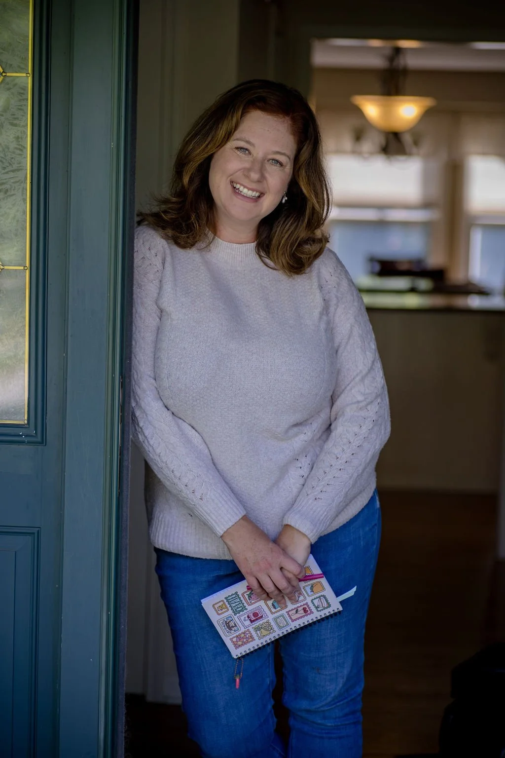 Fiction book coach, Miranda Keskes, has shoulder-length brown hair, is wearing a light sweater and blue jeans, and is standing in a doorway smiling while holding a notebook or planner with colorful designs.