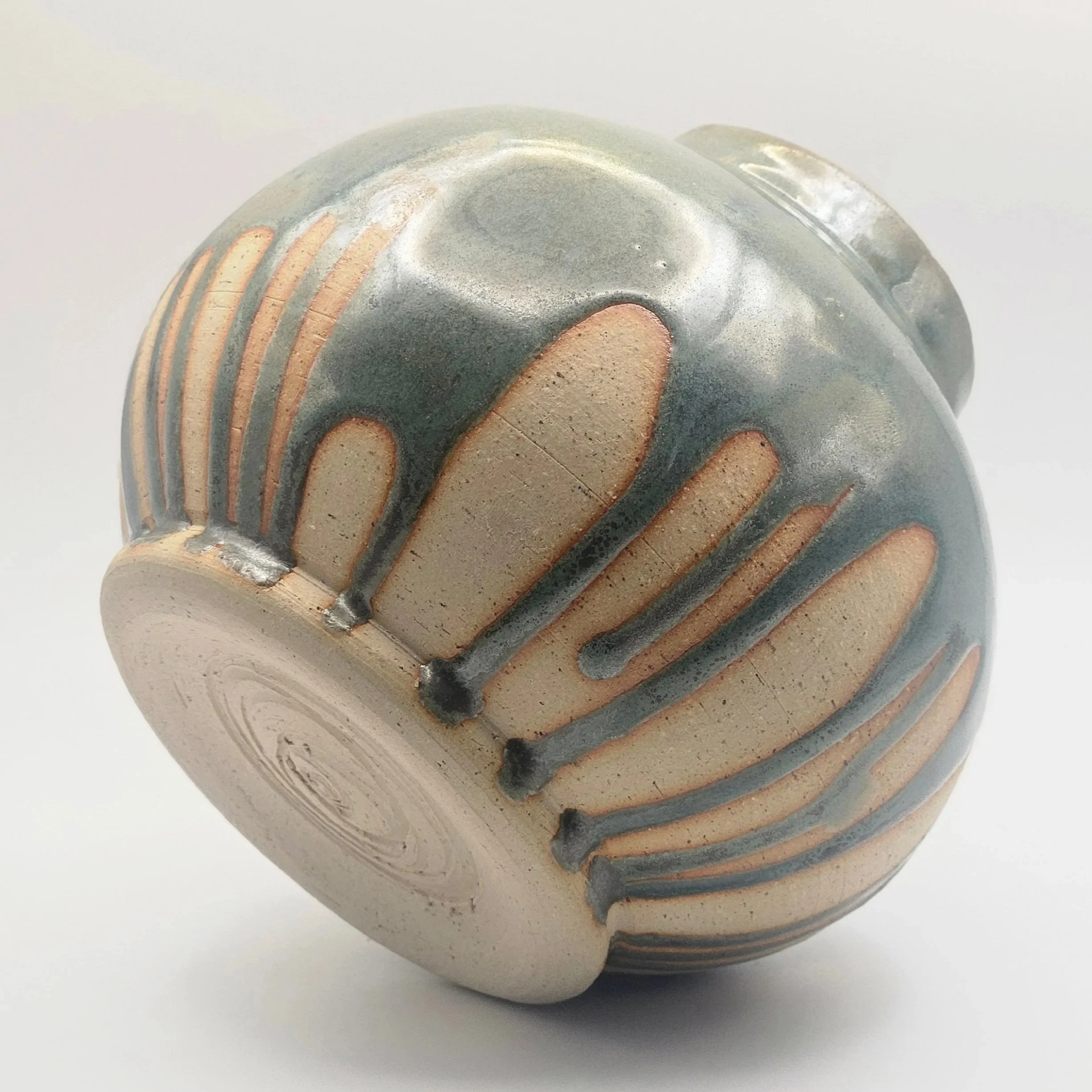 A ceramic or clay vessel with a textured surface featuring vertical ridges and a glossy glaze, photographed from an angle showing its bottom and side.