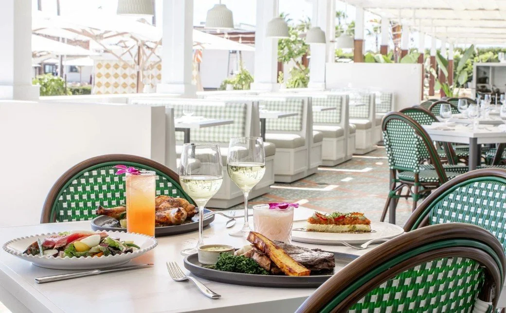 A table set with wine glasses, a salad, grilled meat with vegetables, a chicken dish, and a dessert in a bright restaurant with white walls, green chairs, and outdoor views.