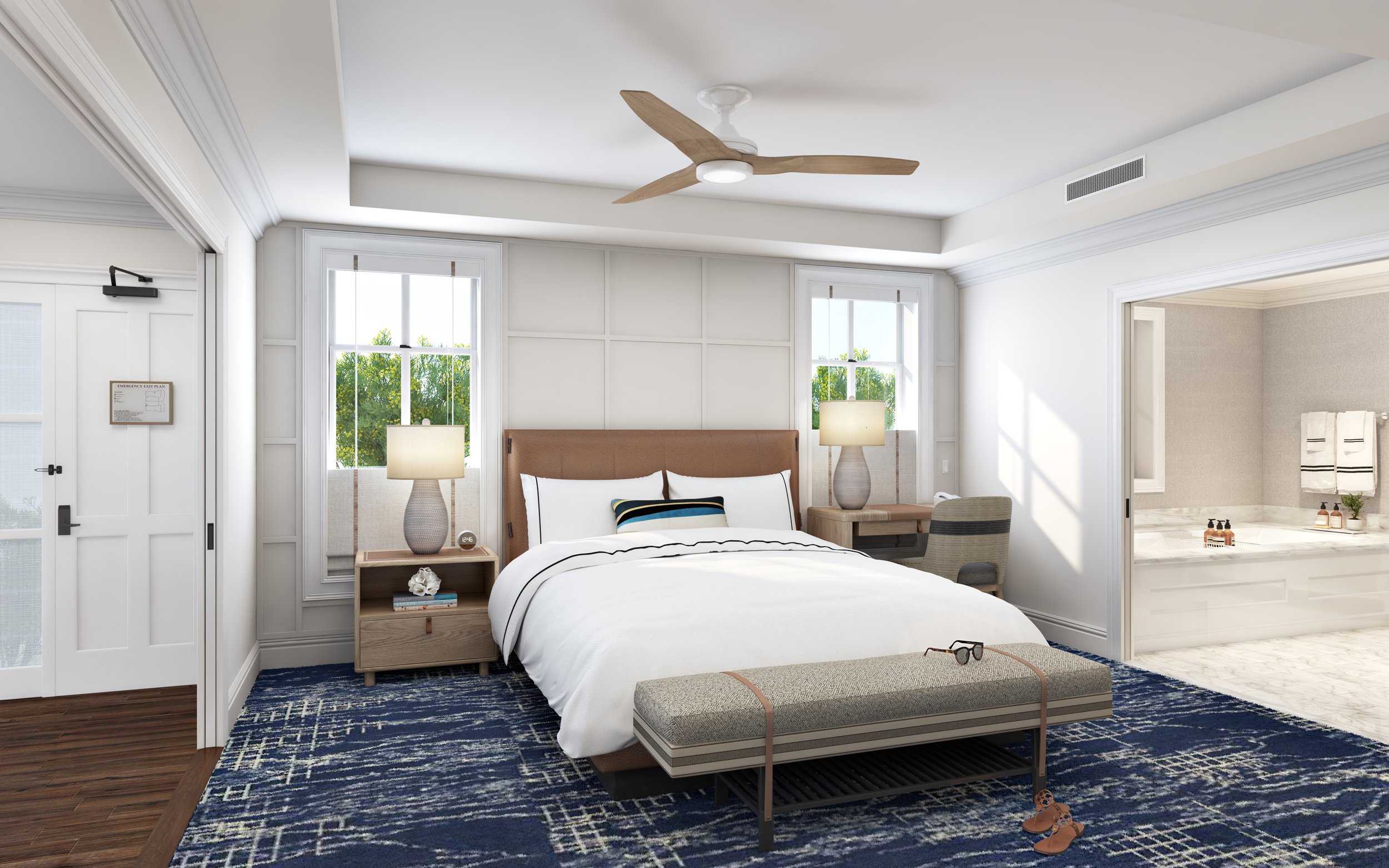 A modern bedroom with a white bed, two bedside tables with lamps, a bench at the foot of the bed with glasses on it, and a ceiling fan. There is a large window behind the bed and an entrance to a bathroom on the right.