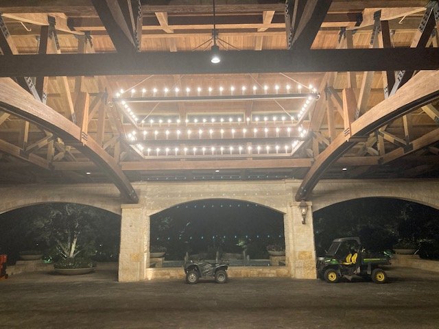 Under a wooden bridge or pavilion with string lights arranged in a rectangular shape hanging from the ceiling. There are arcade or golf cart vehicles parked underneath.