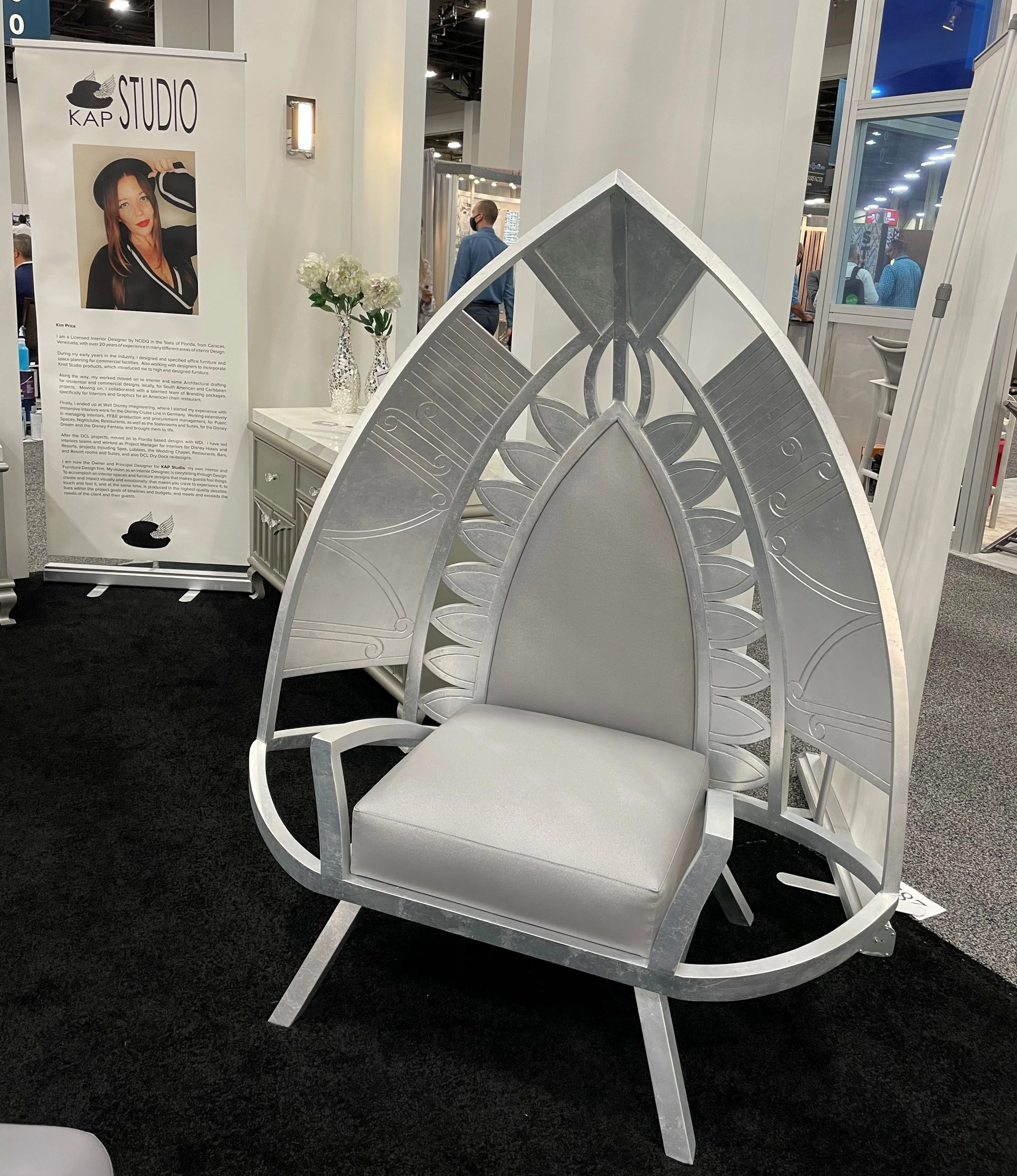 A decorative white chair with an ornate, patterned backrest resembling a peacock feather design, placed on a black carpet at an exhibition or trade show.