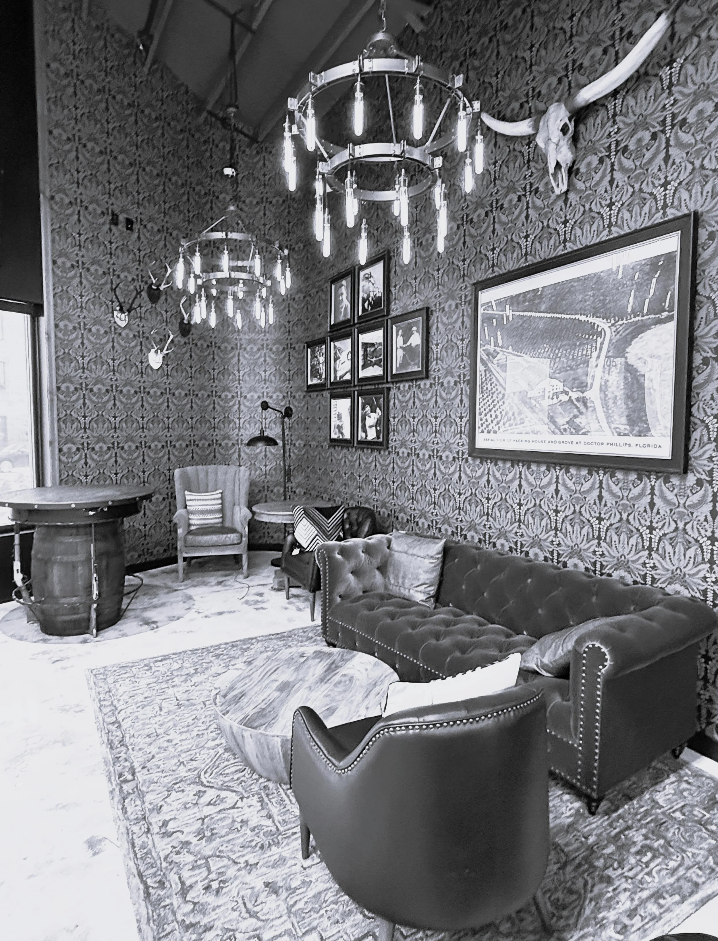 A vintage-style living room with patterned wallpaper, a dark velvet tufted sofa, a leather armchair, a round coffee table, framed pictures on the wall, chandeliers hanging from the ceiling, and animal skulls on the wall.
