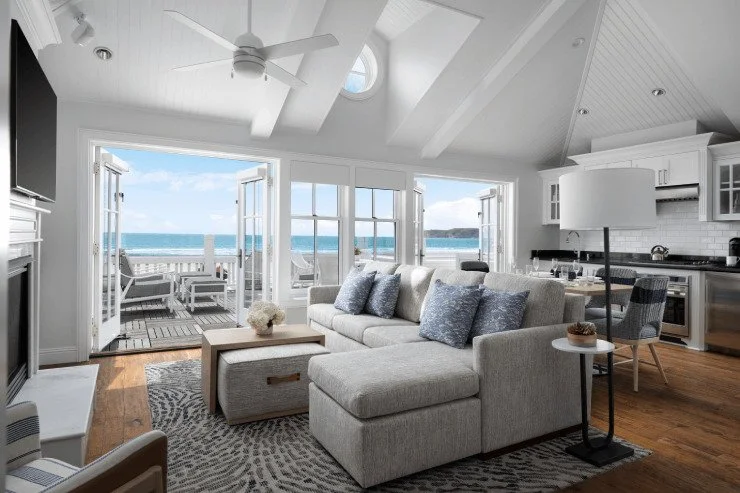 Living room with open back doors leading to a deck overlooking the ocean, with white walls, vaulted ceiling, gray sofa with blue pillows, wooden floor, and kitchen area to the right.