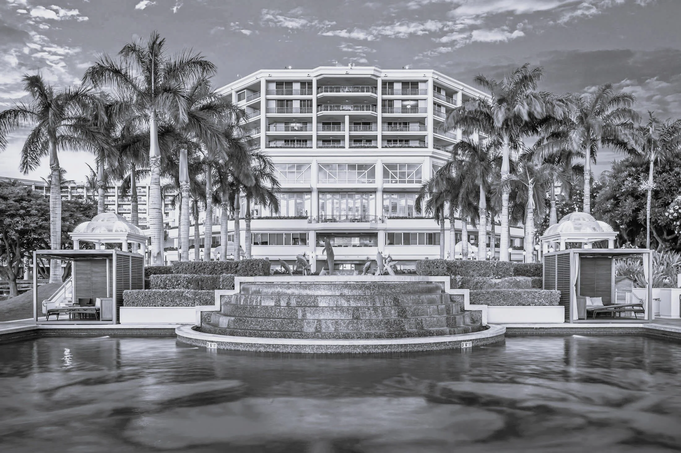 A large hotel with seven floors surrounded by palm trees, with a circular fountain and water feature in the foreground.