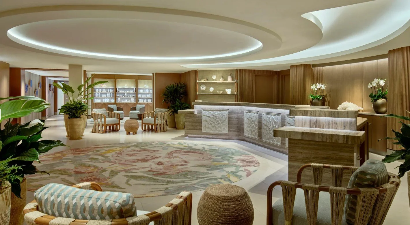 Elegant hotel lobby with a curved reception desk, potted plants, armchairs, and a floral area rug.