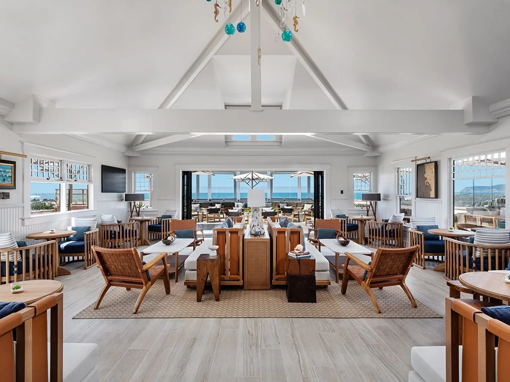 Spacious bright indoor lounge area with large windows, overlooking a patio with umbrellas and an ocean view.