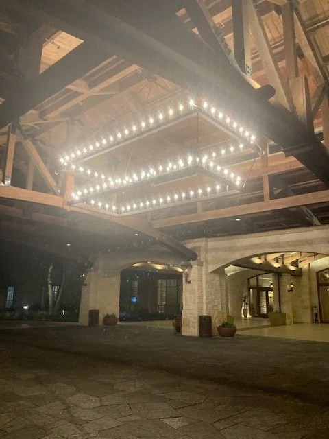 Large square chandelier with multiple small lights hanging in a covered outdoor area of a building.