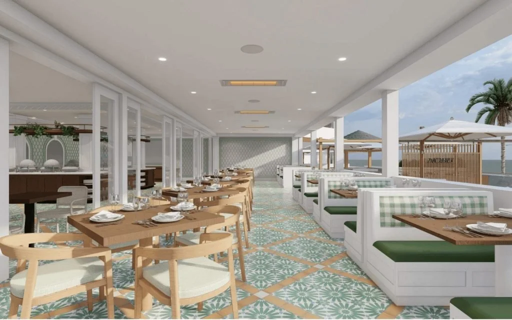 Interior of a modern seaside restaurant with wooden tables, white and green cushioned booths, patterned tile flooring, and outdoor view of the ocean with umbrellas and palm trees.
