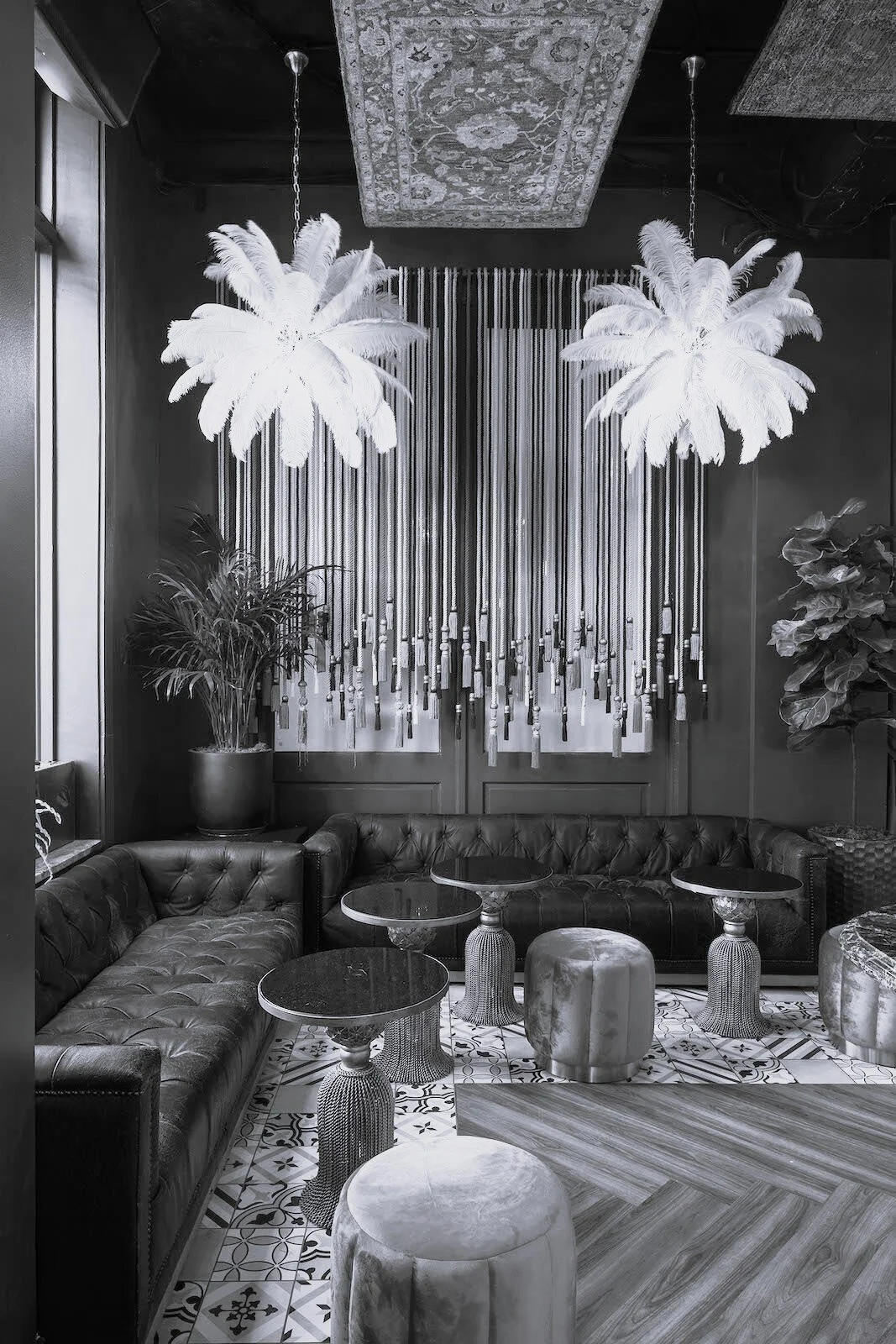 A black and white photo of a luxurious interior lounge area with a curved leather sofa, small round tables with decorative bases, and hanging feather-like light fixtures. There are also large potted plants and patterned floor tiles.