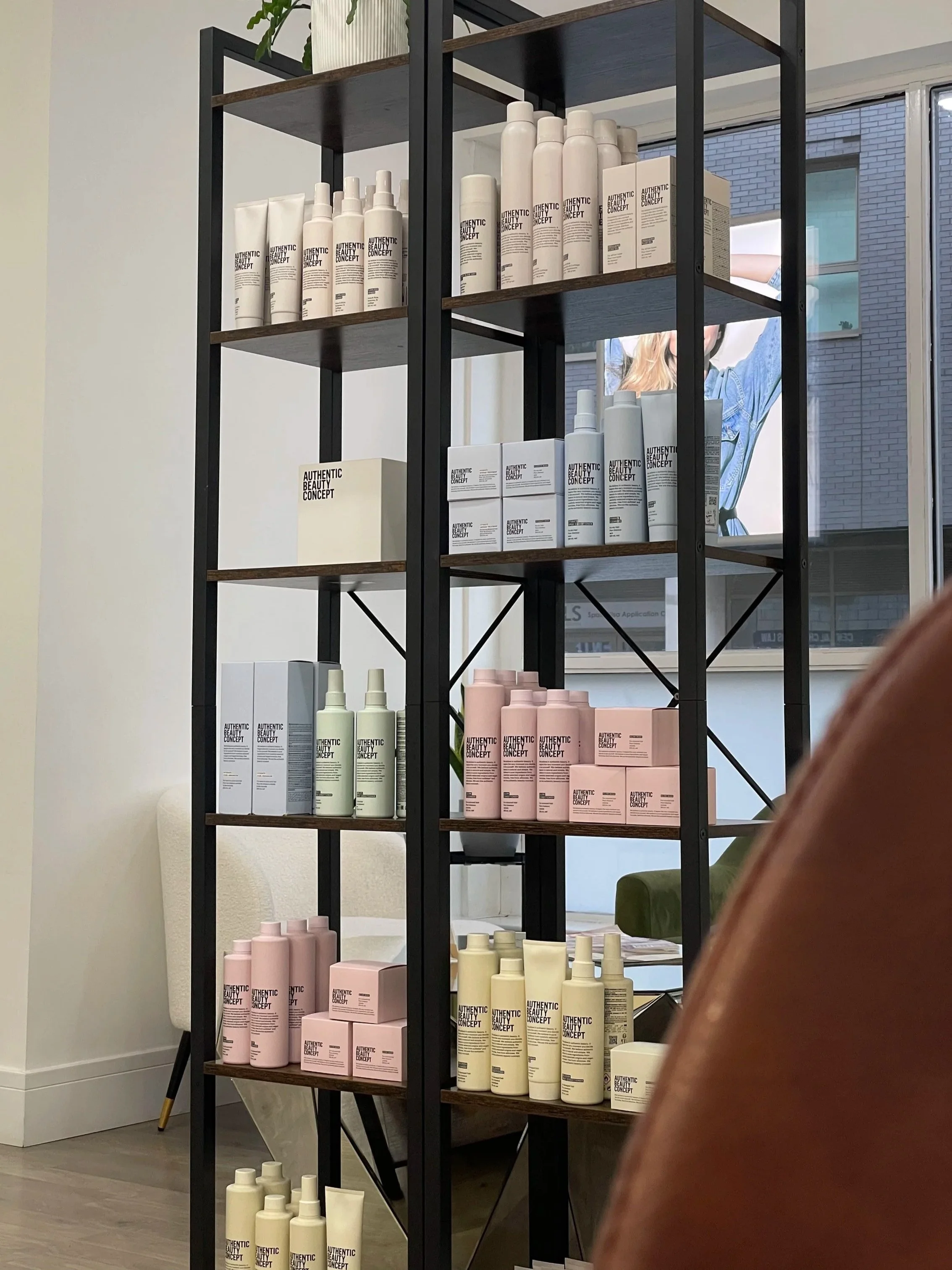 A metal and wood shelving unit displaying skincare or beauty products in pastel pink, white, green, and cream bottles and boxes inside a store or salon, with a window and artwork visible in the background.