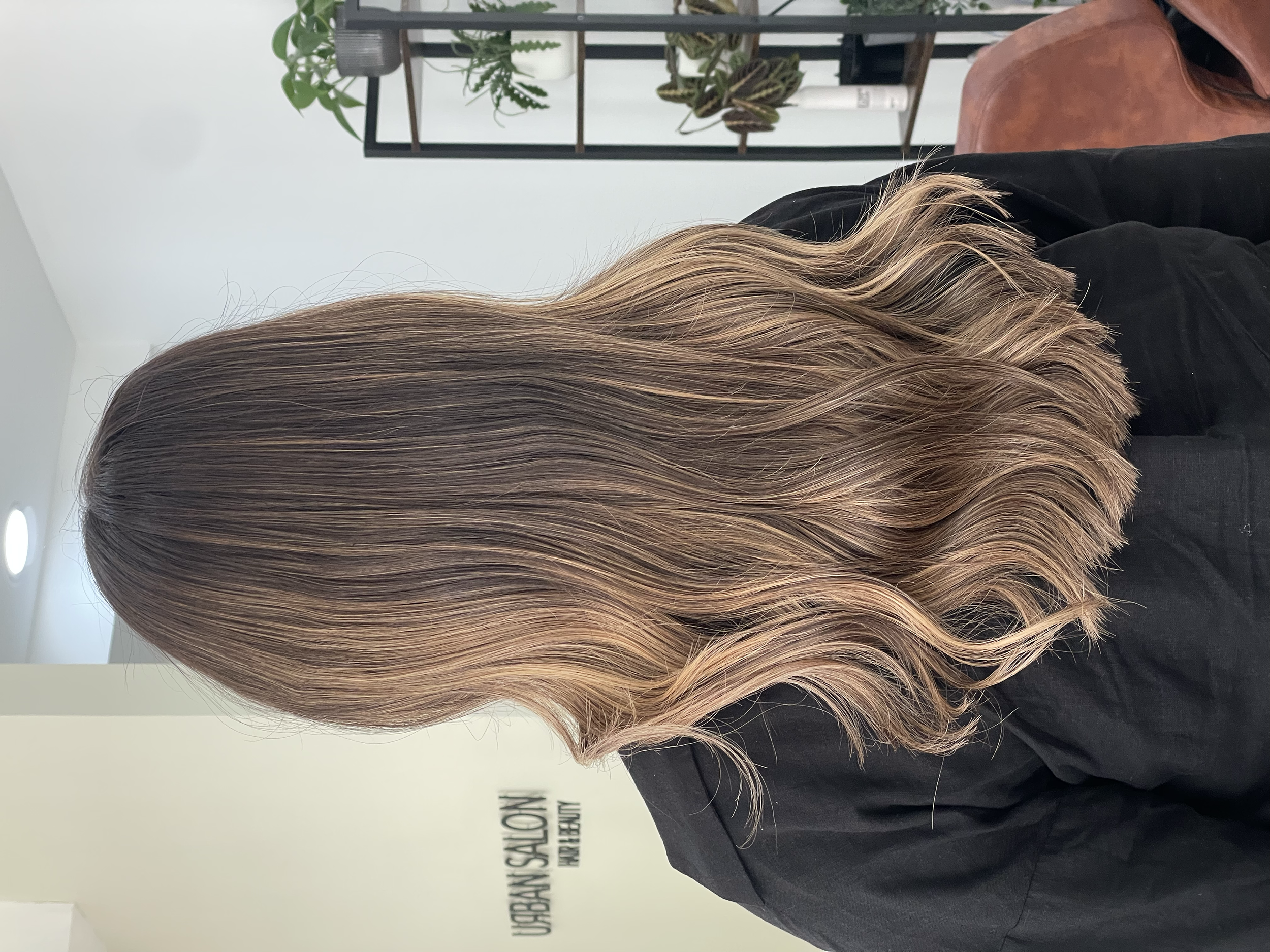 Side view of a person with wavy, light brown hair with highlights, sitting in a salon, with a white wall, shelves with plants, and salon sign in the background.