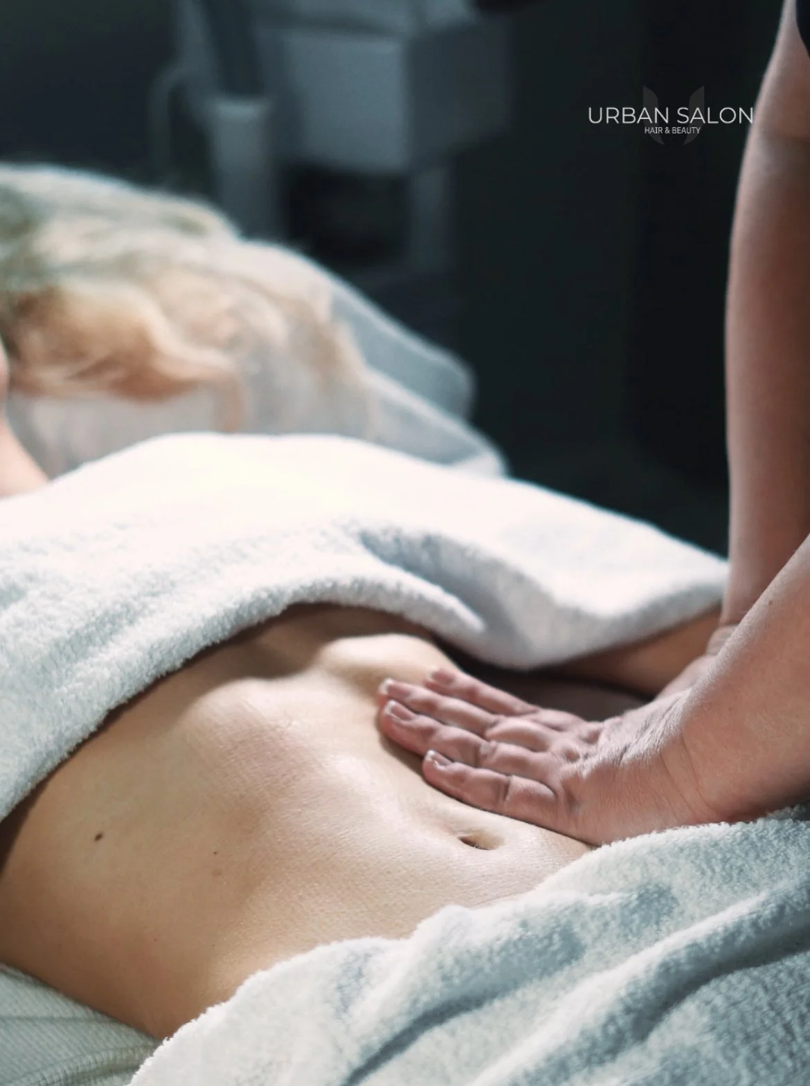 A person receiving a massage on their abdomen at Urban Salon Hair & Beauty, with a towel draped over part of their body.
